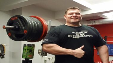 Zydrunas Savickas gives a thumbs up of a approval in front of a weight rack.