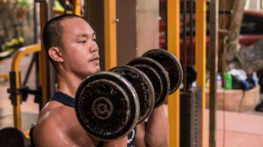 Man sitting in gym holding dumbbells at shoulders