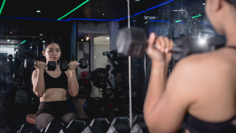 Woman sitting in gym holding dumbbells near shoulders