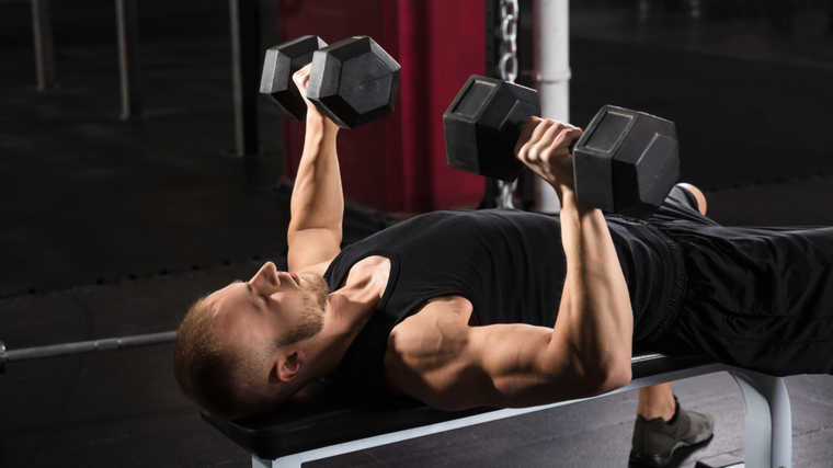 Man in gym lying on flat bench lowering dumbbells