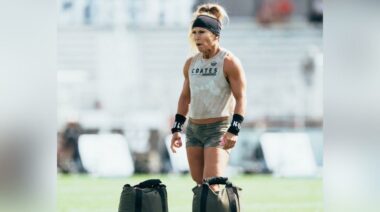 CrossFit competitor wearing a camo tank top and shorts, standing on a turf field in front of two sandbags.